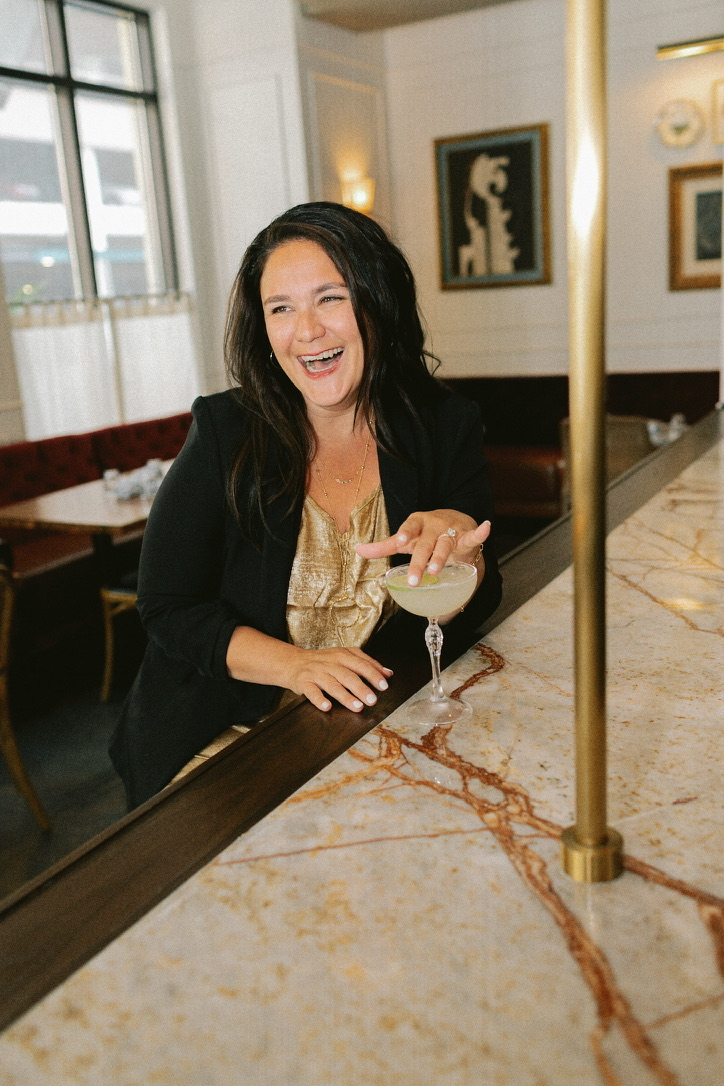 Aly Carter laughing at a marble bar in a gold top and black blazer